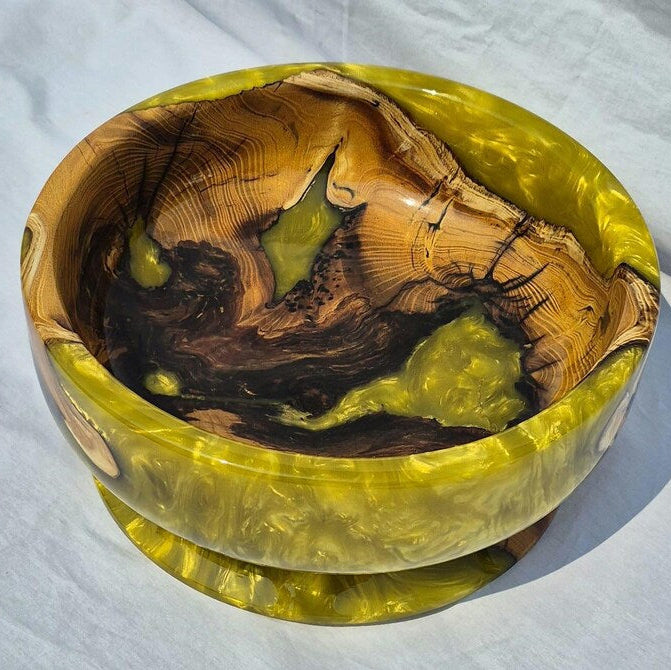 A handcrafted wooden bowl with a glossy, marbled finish featuring swirls of olive green, yellow, and dark brown wood grain, set against a white fabric background.